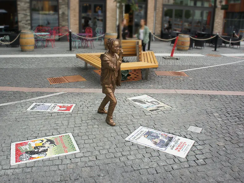 Statyn Ulla vad hände sen? på Ulla Jacobssons plats. En ung flicka som är arg, omgiven av fyra filmaffischer i mosaik, infällda i marken. Konstnär Björn Carnemalm.