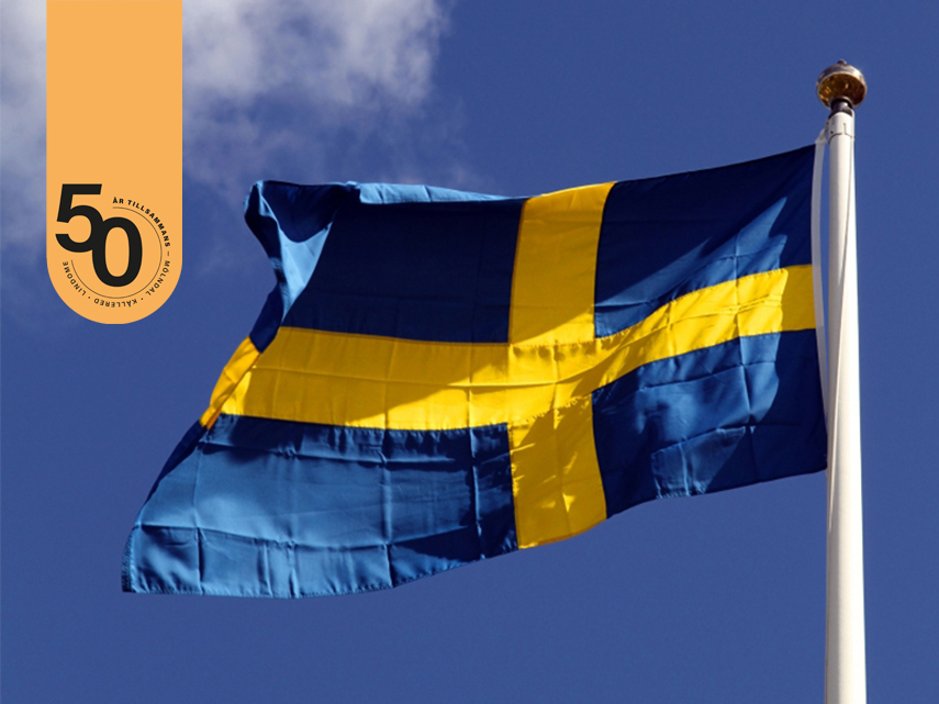 Sveriges flagga mot en klarblå bakgrund. Ett tunt, vitt moln flyter in från vänster. Märke 50-årsjubileum infogat i vänster överkant.