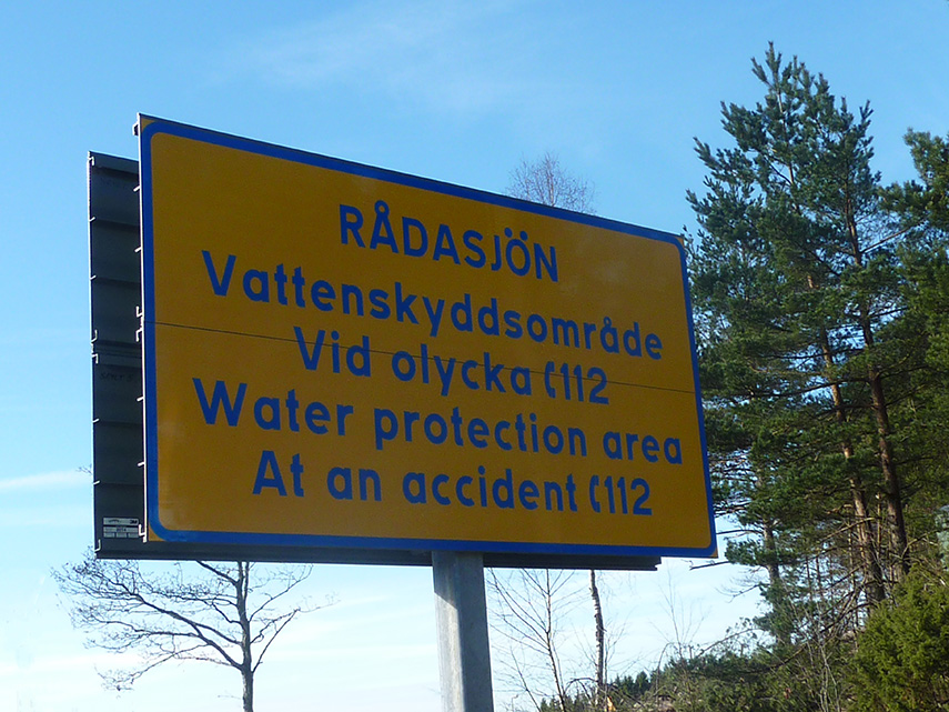 En gul skylt med information om vattenskyddsområde. På skylten står "Rådasjön, Vattenskyddsområde, Vid olycka ring 112. Water protection area, at anaccident call 112".
