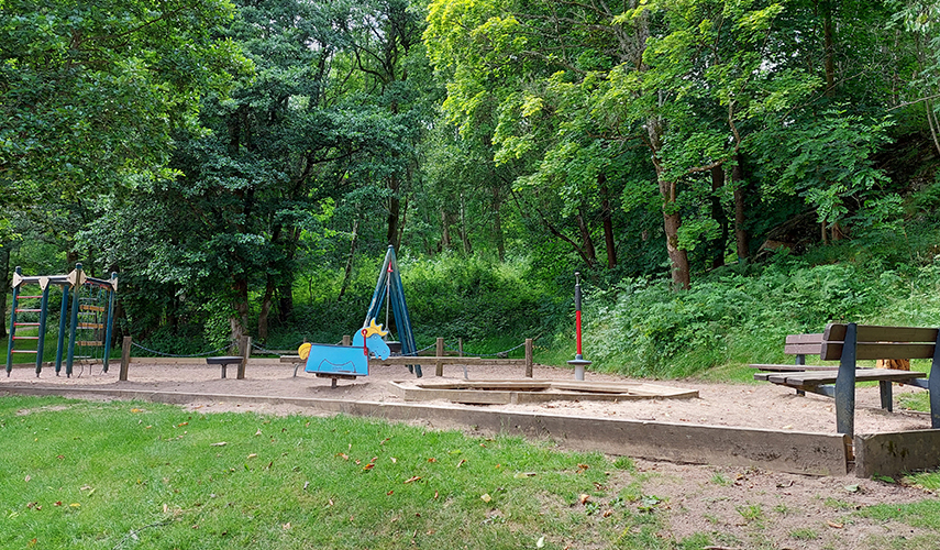 En lekplats med gungdjur, klätterställning, gungor och sittbänkar. Gröna träd står i bakgrunden.