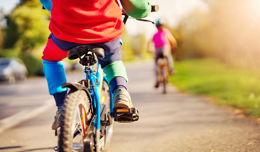 Två barn i färggranna kläder cyklar på en gata. De cyklar bortåt från den som fotar.