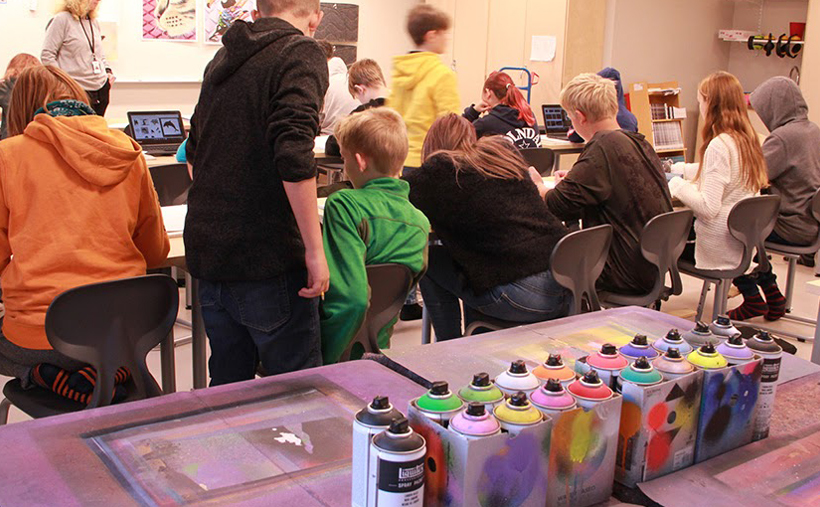 En skolklass i årskurs 6 sitter i klassrummet och förbereder ett street art-projekt. I bildens förgrund syns ett antal färgsprayflaskor i olika färger.