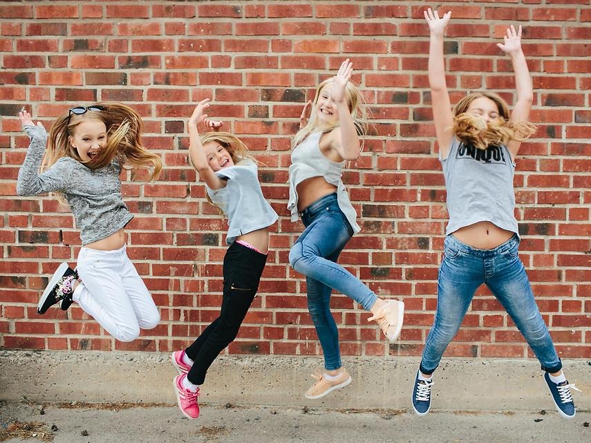 Fyra tjejer hoppar med glädjetjut. Utomhus framför röd tegelvägg