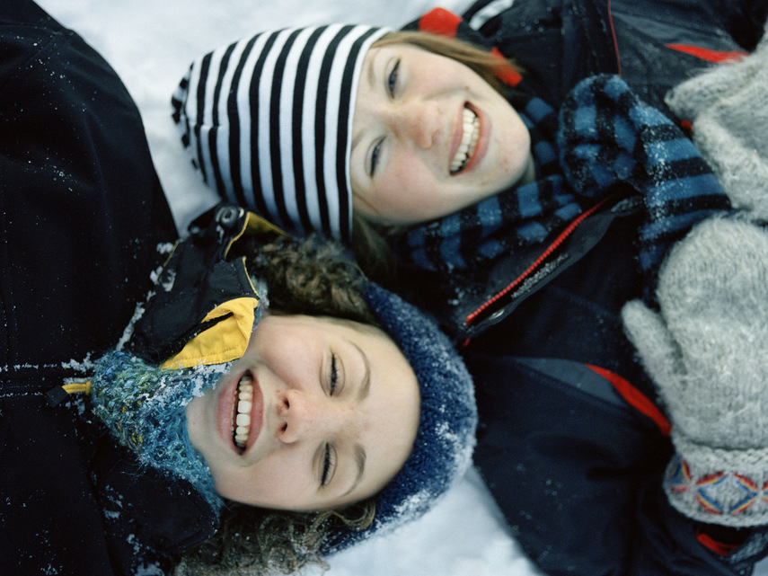 Två  vinterklädda tonårsflickor ligger i snön och skrattar.