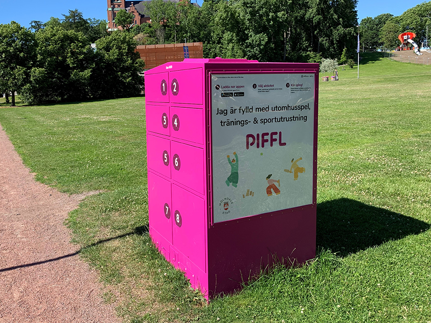 I en park står ett rosa skåp med flera luckor på. På skåpet står en skylt med texten "Jag är fylld med lek, sport och träningsutrustning. Piffl." 