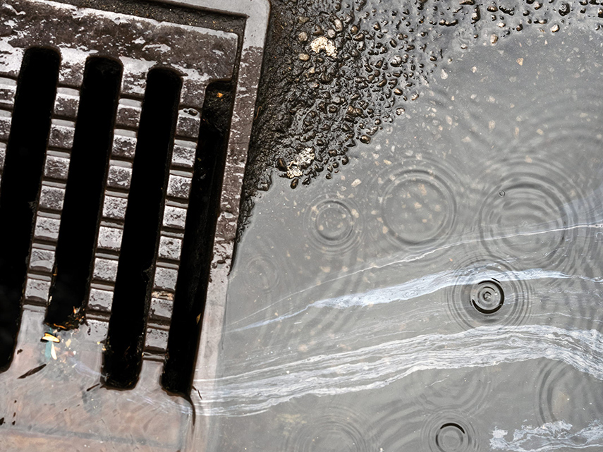 På en gata finns en gallerförsedd dagvattenbrunn. Regn faller ner på asfalten och bildar krusningar på vatten som rinner ner i brunnen