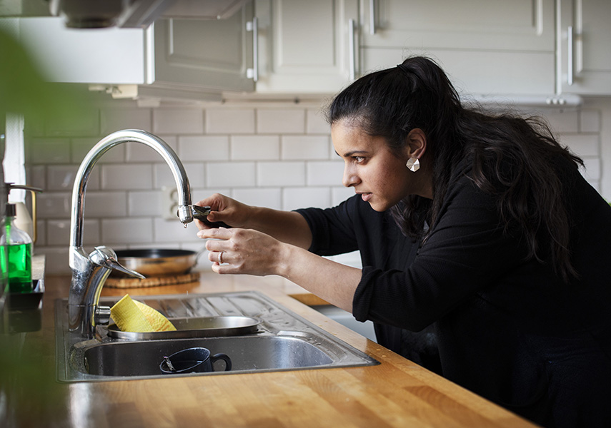 En kvinna står vid sin köksbänk. I höger hand håller hon en skiftnyckel som hon skruvar åt silen på vattenkranen med så den inte ska droppa.