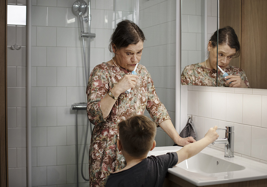 En kvinna står i badrummet och borstar tänderna. Hennes son kommer in och stänger av vattenkranen som inte behöver vara på när hon borstar.