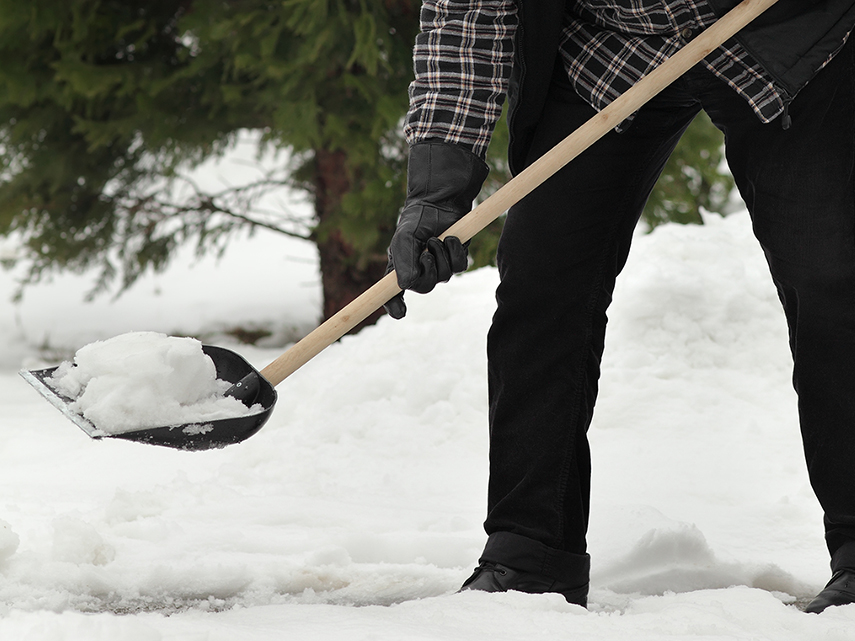 En person står ute i snö och håller i en snöskyffel. Med skyffeln skottar han trottoaren.