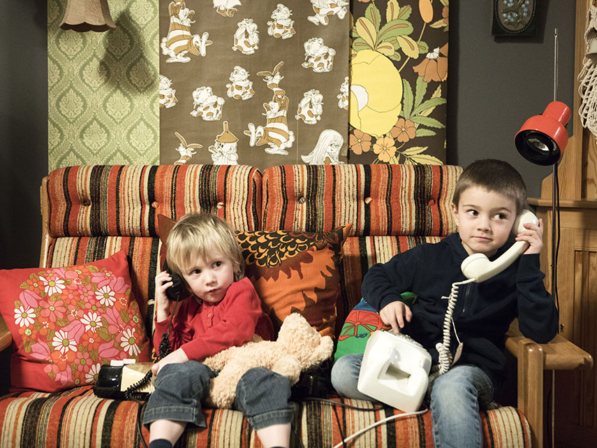 Två små barn sitter i en 70-talssoffa med varsin gammal telefon.