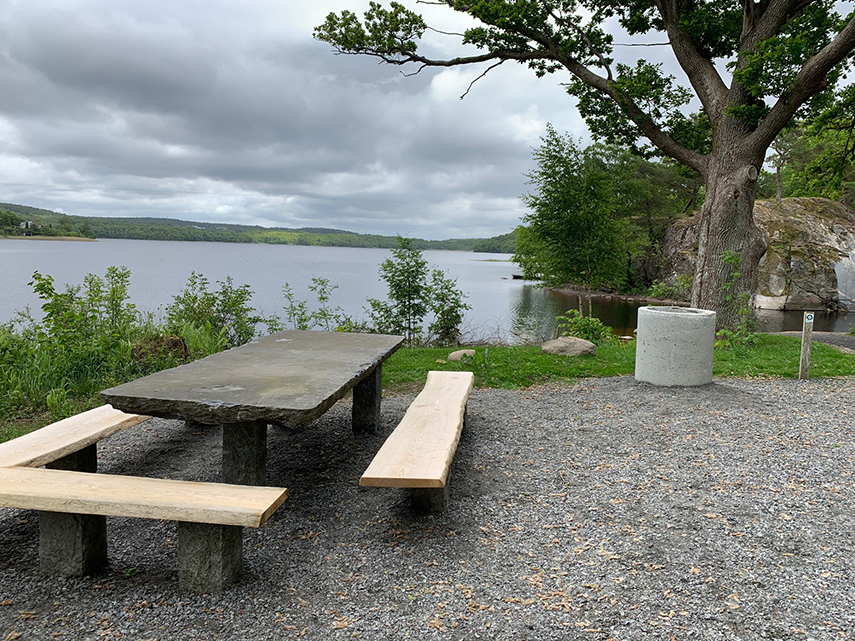 Ett bord i sten med bänkar i ekträ står vid en strand. Till höger finns en liten grillplats.