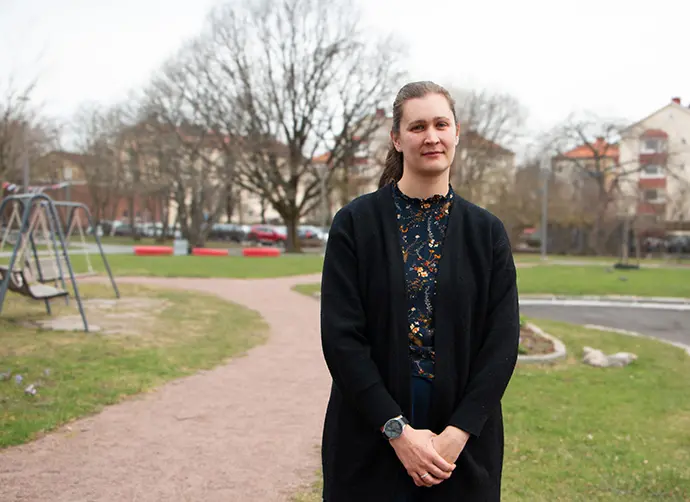 En kvinna står framför en park med en lekplats i bakgrunden. Personen bär en mörk cardigan och en mörk blus med mönster.