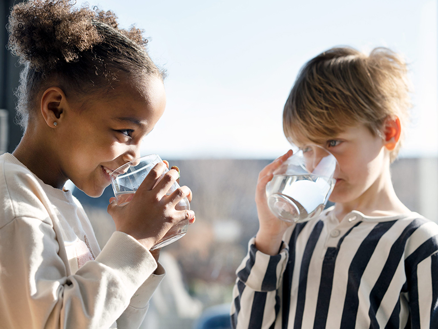En flicka med ljus tröja och en pojke med en randig tröja står och tittar på varandra samtidigt som de dricker vatten ur varsitt glas.