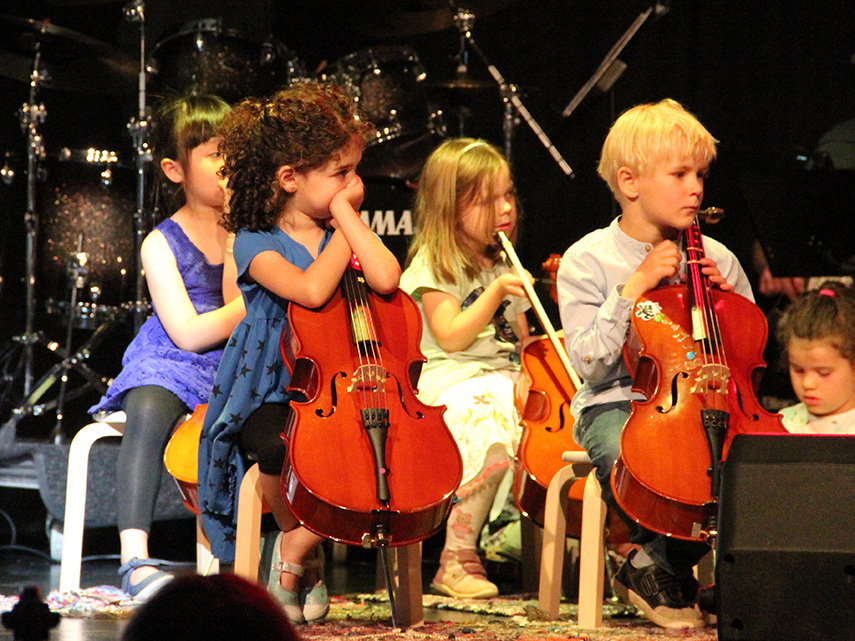 Fyra fundersamma barn sitter på varsin pall och lutar armarna mot varsin cello. Foto: Victoria Eriksson, 2018