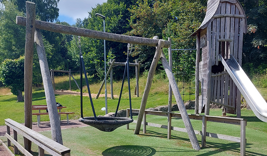 En lekplats med utrustning i trä. Gunga, klätterhus, rutschkana och linbana.