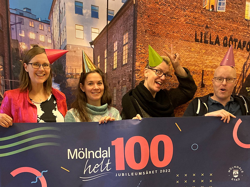 Foto på fyra personer bakom en banderoll med loggan för Mölndals 100-årsjubileum. 