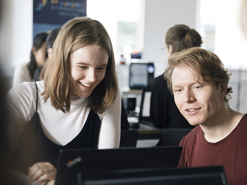 En manlig elev med keps jobbar fokuserat vid datorn. I bakgrunden ser man en annan elev som får hjälp av en lärare.