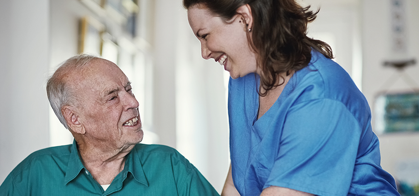 En äldre man tittar upp mot en kvinnlig undersköterska som ler mot honom.