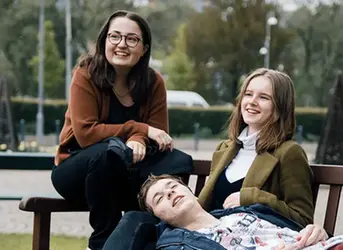 Tre elever sitter tillsammans på en bänk utomhus och umgås i en avslappnad miljö.