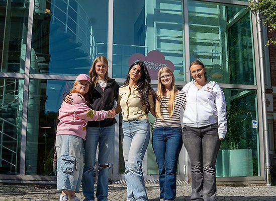 Fem elever står utanför skola och poserar. ler.