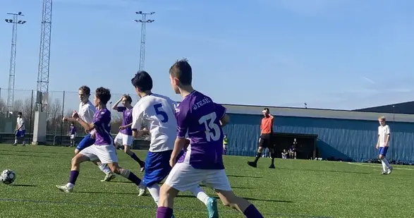 Fotbollsspelare i vita och lila tröjor jagar en boll på en fotbollsplan.