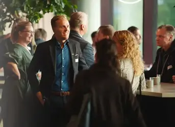 En grupp personer i kostymer och kavajer samlas vid ett bord med kaffemuggar. En man bär en blå skjorta och ett visitkort med texten "‘Networking’".