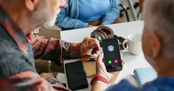 En person pekar på en smartphone som ringer. En mugg och hörlurar syns på bordet.